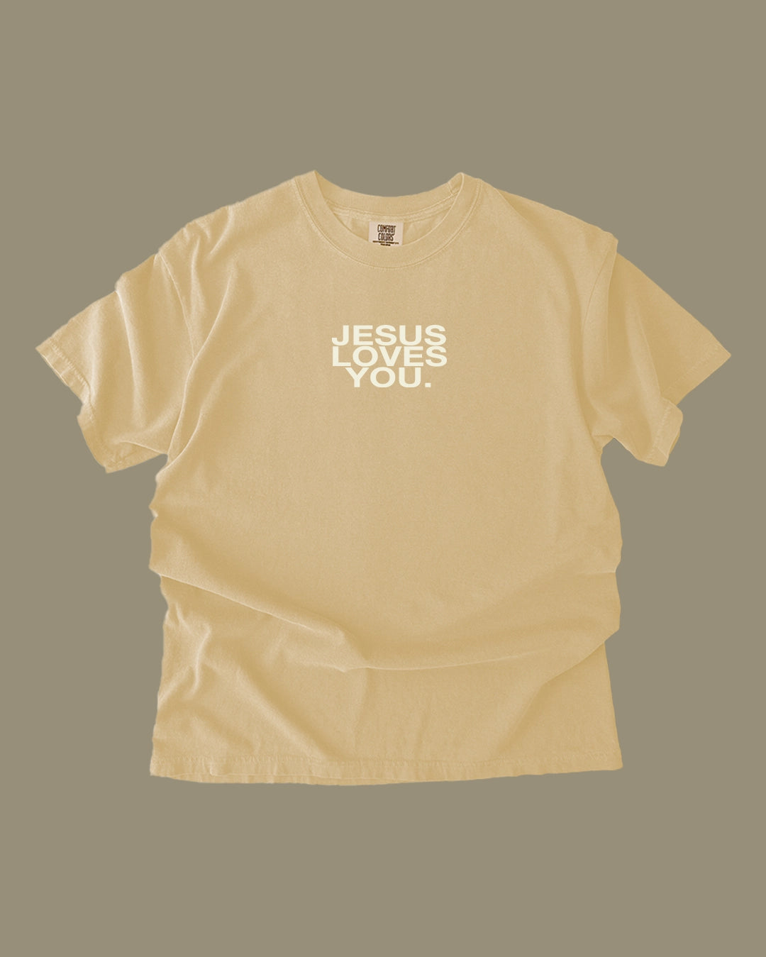 A pale yellow t-shirt with the text 'JESUS LOVES YOU.' printed in white block letters on the chest, displayed against a solid taupe background.